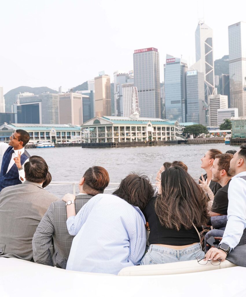 一群人在維多利亞港的遊艇上欣賞香港天際線,背景可見中環高樓大廈與海濱建築,氣氛愉快放鬆,展現專屬遊艇體驗的奢華與活力。